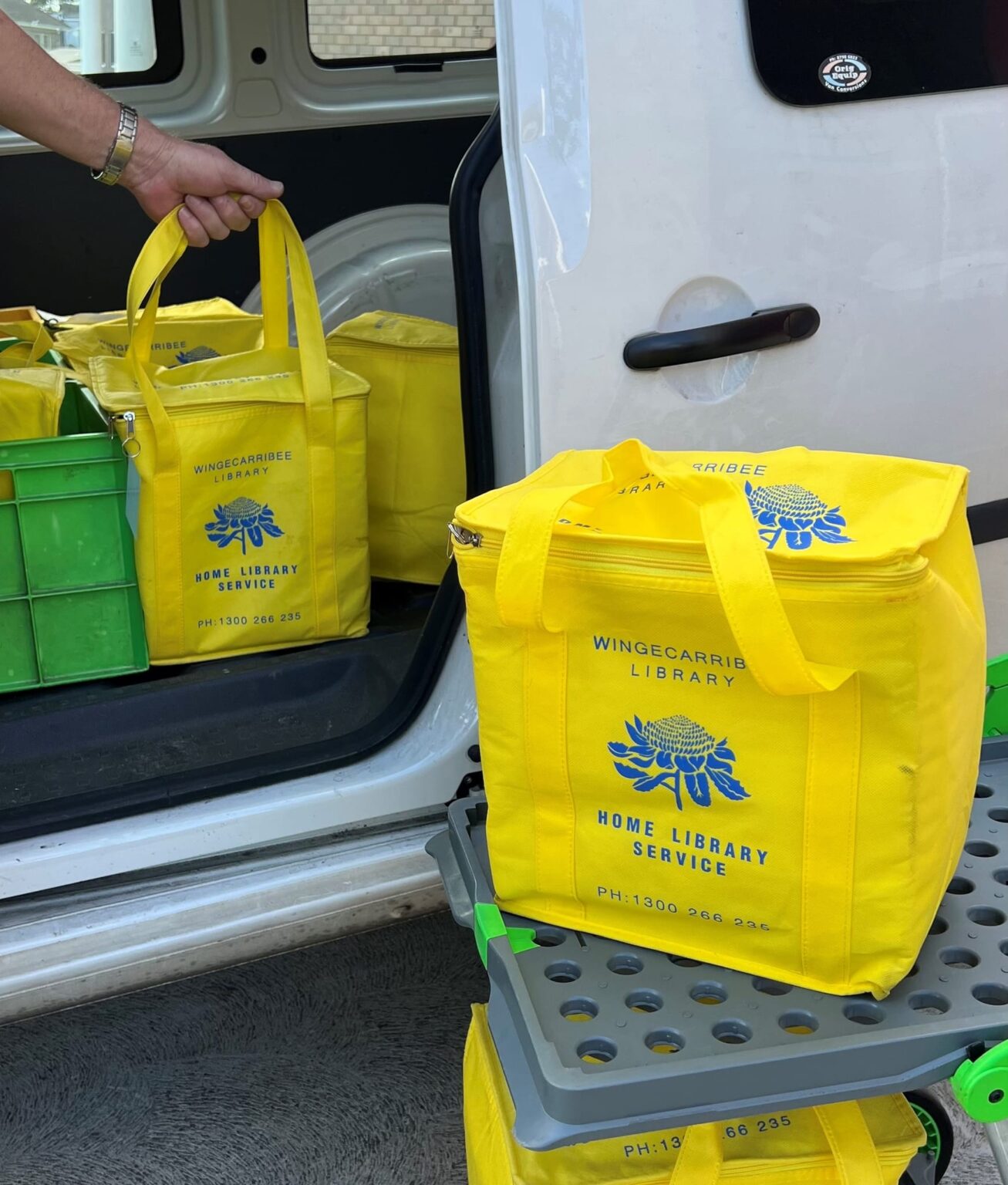 Yellow Home Library Service bags being loaded into a van sliding door. A Yellow bag sits on a trolley waiting to be loaded.