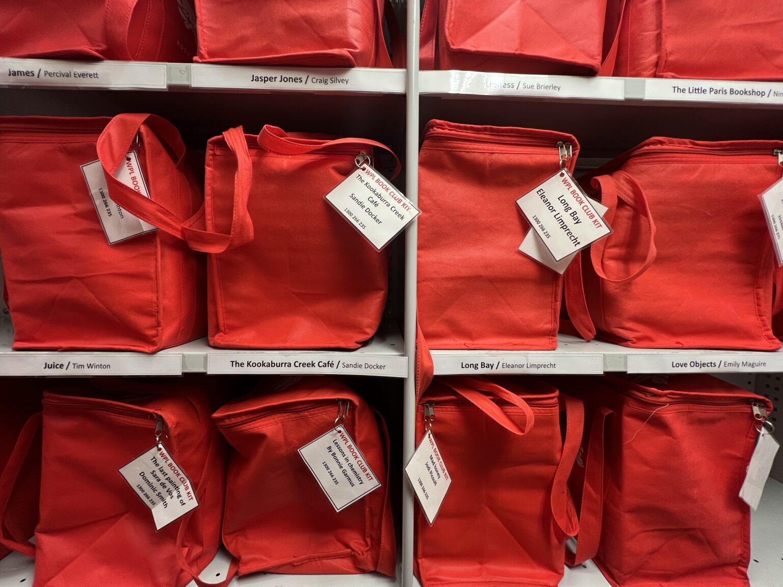 12 red book club kit bags sit on 3 rows of shelving. There are 4 book club kit bags in each row. Laminated white tags show the book title and author of each kit.