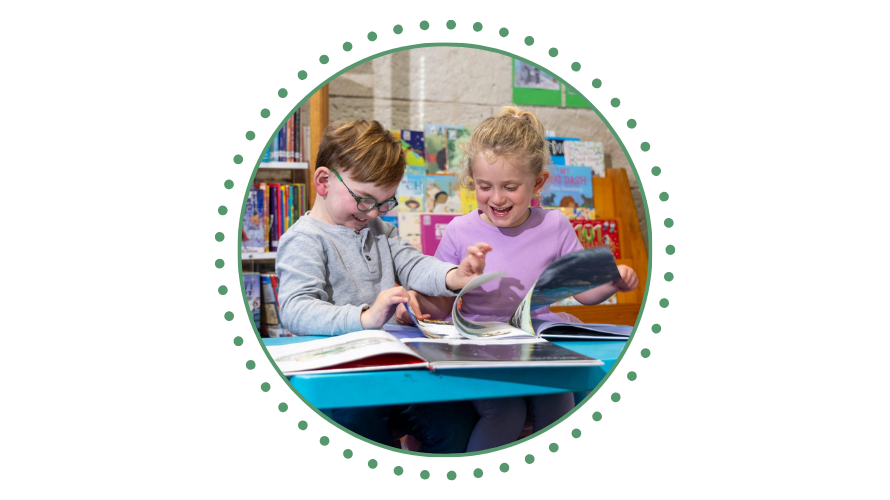Two children happily reading books at a table in Mittagong Library with picture book shelving in the background.