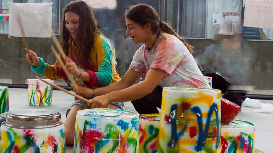 Two teenagers playing drums made out of buckets, splattered in paint.