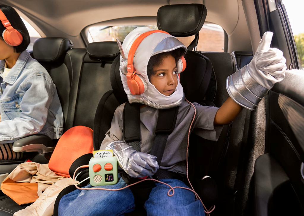 A boy in a car dressed up as an astronaut listening to his Yoto mini player. He is gesturing a thumbs up out the window as he listens to the Yoto.